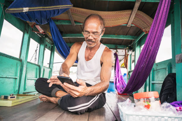 vietnamese senior man looking at mobile on vessel at floating market in mekong delta vietnamese senior man sitting at boat deck and looking at mobile on floating market in Can Tho, south vietnam vietnamese old man portrait stock pictures, royalty-free photos & images
