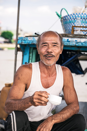 vietnamese senior man holding tea cup and smiling to camera on vessel at floating market in mekong delta in Can Tho, south vietnam
