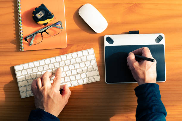 In a zenithal view. A man's hands on a keyboard and a graphic card. The desk is a table at home, he is teleworking.