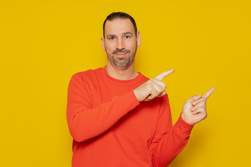 Smiling happy man 40s in casual red pullover pointing index fingers to the side in mock-up workspace area isolated on yellow colored background studio portrait. People lifestyle concept.