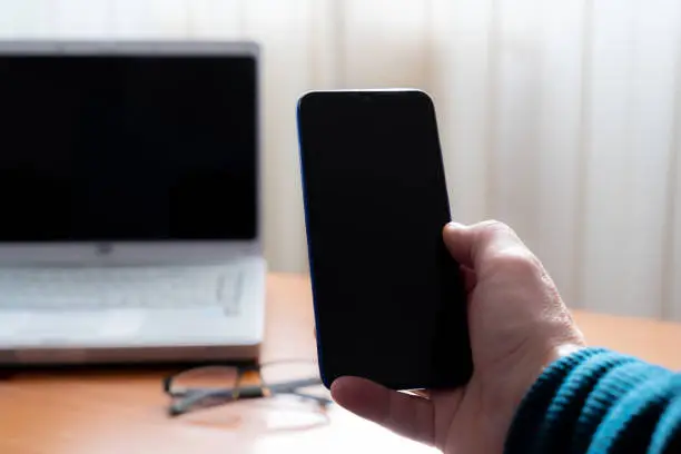 A human hand holding a smartphone in front of a computer. It is the inside of a home, teleworking.