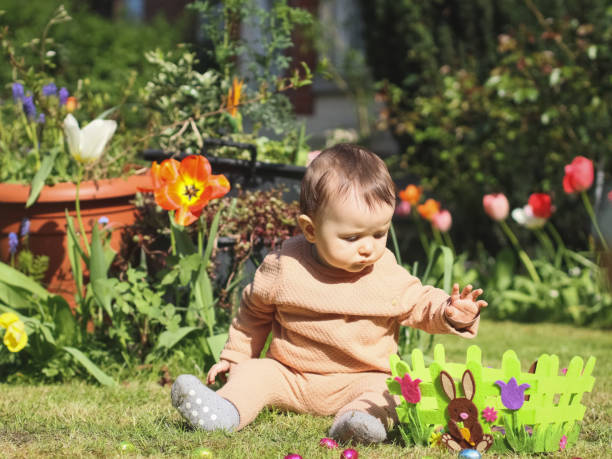 小さなかわいい白人の女の赤ちゃんが花の間の芝生に座って、チョコレートのフェルトイースターバスケットのハンドルで手を伸ばします - baby holding babies only sign ストックフォトと画像