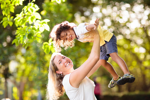 Mother and son in sunny park. Loving interracial family playing outdoor. Mom holding little boy high in the air. Young woman and adorable child play and laugh. Happiness and love. Happy mothers day.