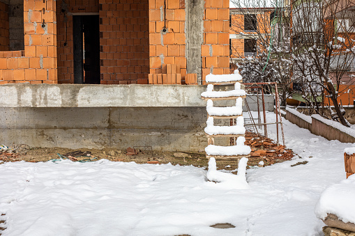 Wooden staircase and building under construction. There is snow on the top of the ladder. The ground is snowy. The building is under construction. We see concrete and bricks.