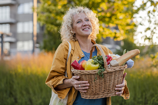 A happy mature woman is coming back from Farmer's market. She is shopping with an eco-friendly shopping basket for a sustainable lifestyle.