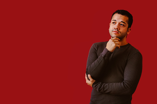 Portrait of serious young trans masculine man wearing dark gray top with hand on chin, thinking, against red background