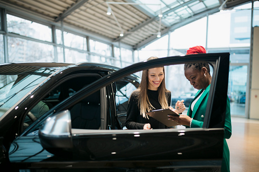 Saleswoman and a female customer in a car dealership. Sales manager explaining looking at clipboard and explaining the car features to woman customer in showroom.