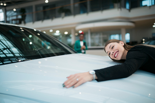 Beautiful woman hugging and showing her love to a car at auto dealership. Happy female choosing her dream car to buy at showroom.