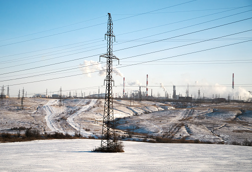 Big factory with many pipes with smoke in the clear blue sky.\nElectricity Pylon power line transmission tower in front