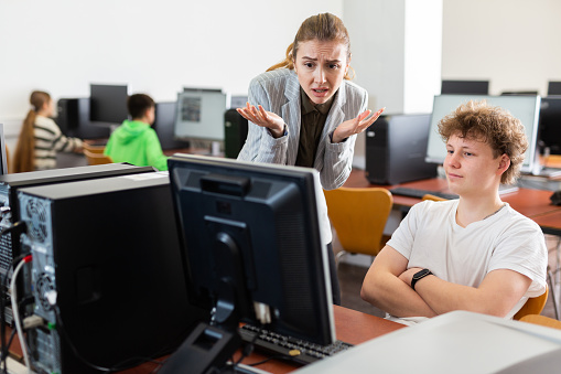 Frustrated worried young female teacher emotionally talking to neglectful teenage student sitting at computer in classroom during lesson