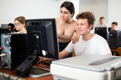 Friendly teen schoolgirl helping interested focused male classmate sitting at computer in information technology class of school