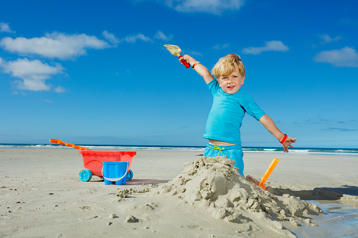 Happy cute little blond handsome boy play in sand with shovel lifting hands up on ocean summer vacation