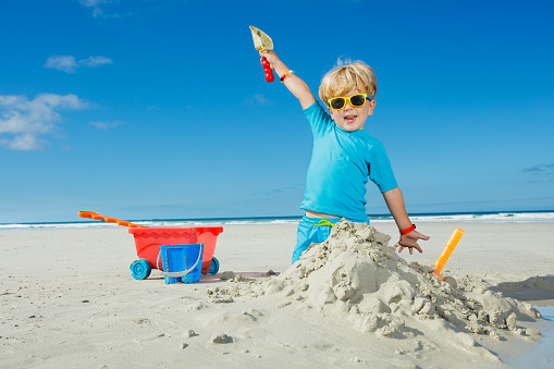 Happy cute little blond handsome boy play in sand with shovel lifting hands up wearing sunglasses on ocean summer vacation