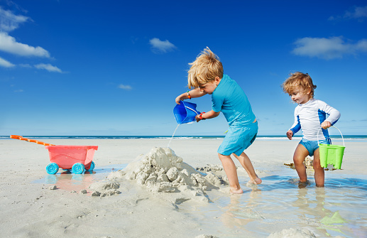 Cute little blond boy play with sand pouring water from plastic bucket on the sea beach during vacation