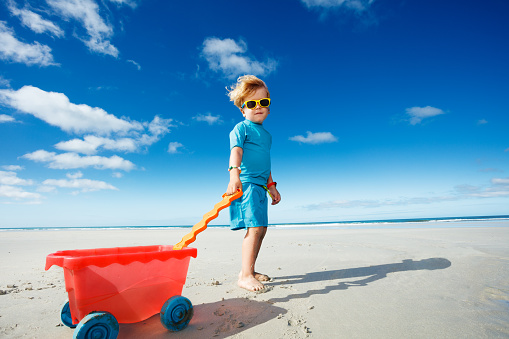 Happy cute blond boy in sunglasses walk on the white sand beach playing with toy cart looking back