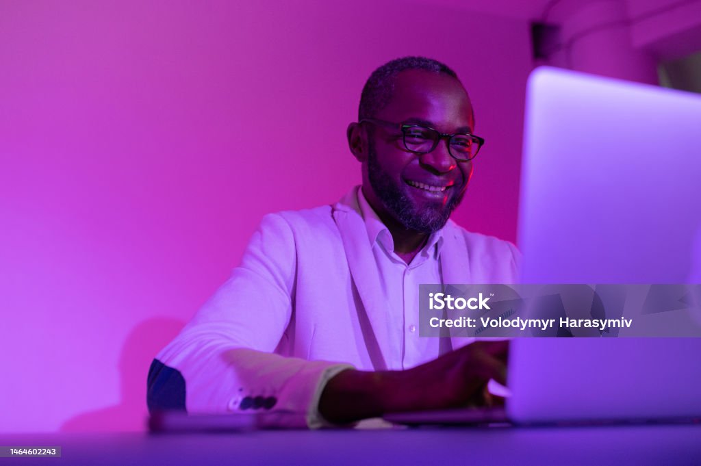 a black man in a white suit and glasses works at a laptop. a man in glasses works in the office in the evening a black man in a white suit and glasses works at a laptop. a man in glasses works in the office in the night Purple Stock Photo
