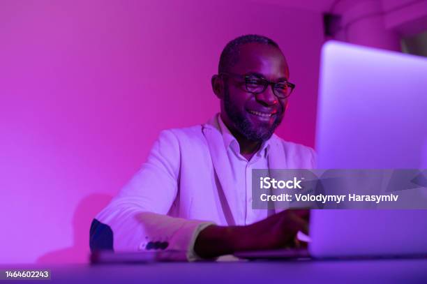 A Black Man In A White Suit And Glasses Works At A Laptop A Man In Glasses Works In The Office In The Evening Stock Photo - Download Image Now