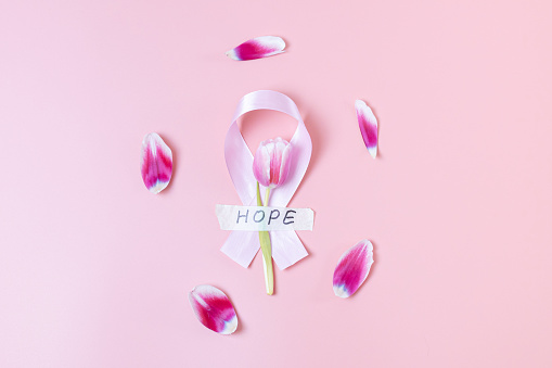 One wide satin pink ribbon with a tulip, sticky tape with the word: hope and petals scattered around lie in the center on a pink background, flat lay close-up. World cancer day concept, breast cancer.