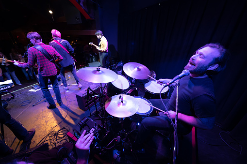 An Americana folk-rock band performing onstage in front of an audience at a live music venue. A videographer is filming the performance.