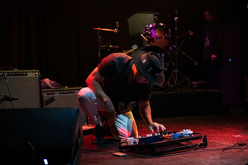 A young Latino man wearing a wide brim hat kneeling down and adjusting a deck of guitar effects pedals on a stage.