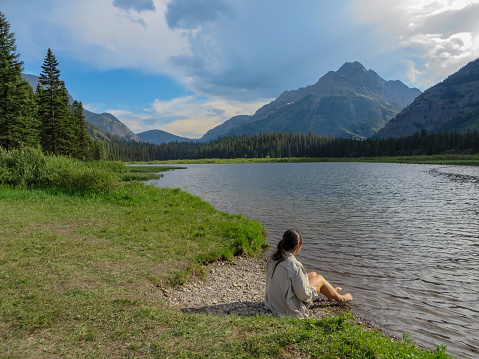 Woman sits with bare feet next to Kootenai Lakes in Glacier National Park with Mountains and Storm Clouds