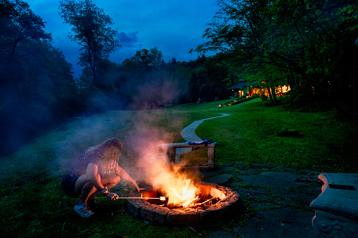 Woman sits roasting marshmallows in a firepit near a river at night, Farmington, Pennsylvania, USA