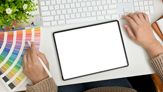 Top view of a graphic designer office desk with digital tablet white screen mockup, color palette, keyboard computer and decor. A female graphic designer working at her desk