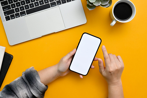 A female hand holding a smartphone white screen mockup, using her mobile phone in modern trendy workspace with yellow tabletop background. Workspace concept