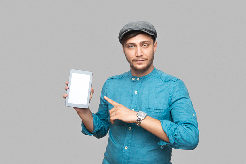 Studio portrait of a smiling young man showing a digital tablet with a blank screen over gray background. Horizontal shot.