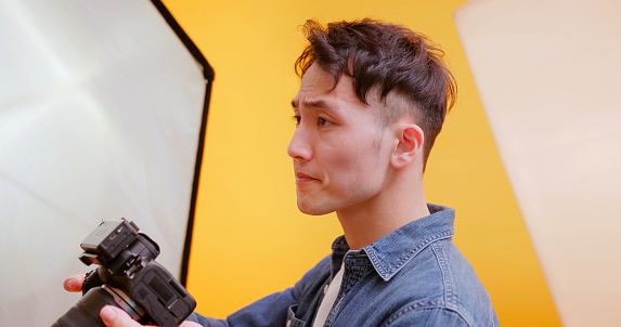 asian young male photographer holding professional camera for shooting standing in front of yellow background at studio - he is  frustrated