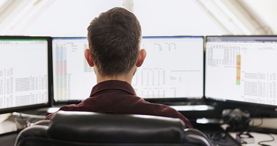 Rear view of a young businessman working on a computer with multiple monitors at his desk in a modern office