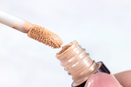 Beige skin concealer pouring from a brush into the bottle on light background close up.