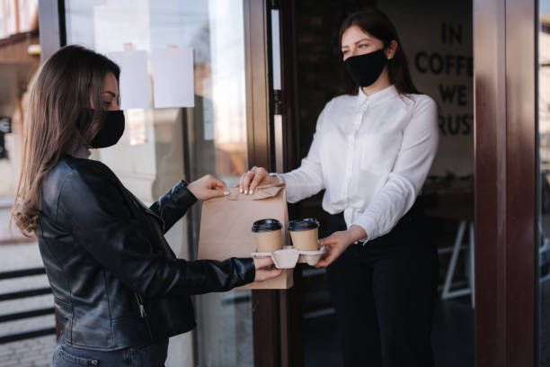 istock Young woman standing outdoors in mask and paying bill through smartphone using NFC technology in a cafe. Female customer paying using contactless technology. Woman taking at order. Take away food concept 1464674941