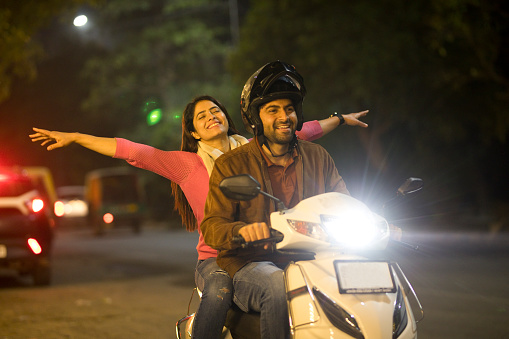 Carefree woman having fun with arms outstretched on motor scooter seat behind her boyfriend at night