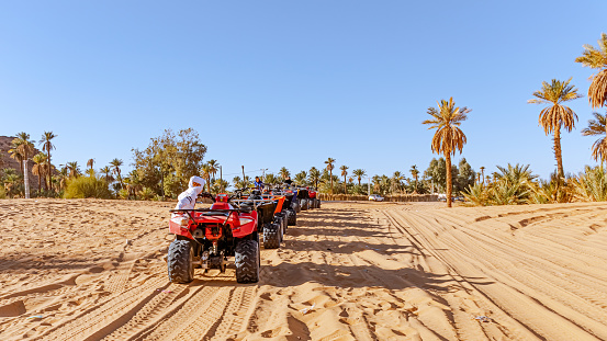 Taghit, Bechar/Algeria - December 27, 2022: Sunny day with sand, palm trees and unrecognizable tourists standing near the queued quad bikes.