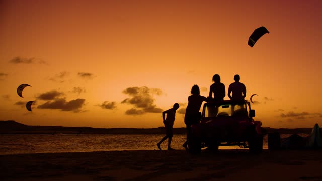 Silhouette of friends group sitting on car and watching people kitesurfing at sunset