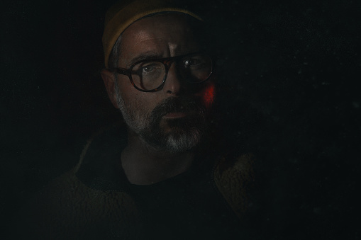 A middle aged Spanish man in eyeglasses looking with a serious facial expression in the darkness