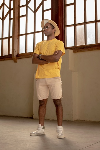 A vertical shot of an Afro-American young male in a yellow t-shirt and straw hat posing with his hands crossed