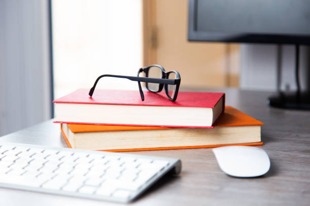 A computer desk with books, glasses, keyboard, and mouse on it