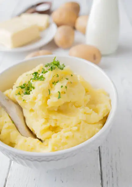 Homemade mashed potatoes or potato puree. Delicious gluten free side dish for dinner or lunch. Served in white bowl with wooden spoon on white wooden background with ingredients. Closeup and vertical image