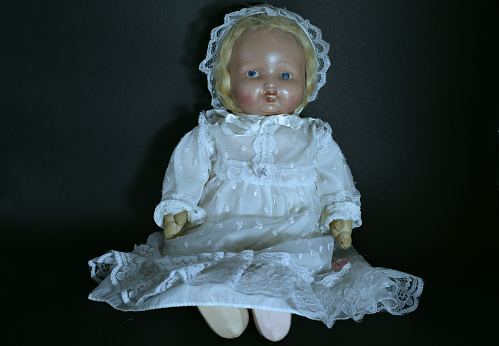 A closeup of a creepy old doll in a white dress on black background