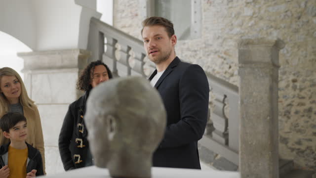 Attractive Adult Caucasian Male Museum Employee Presenting A Statue In A Museum To A Diverse Group Of Tourists