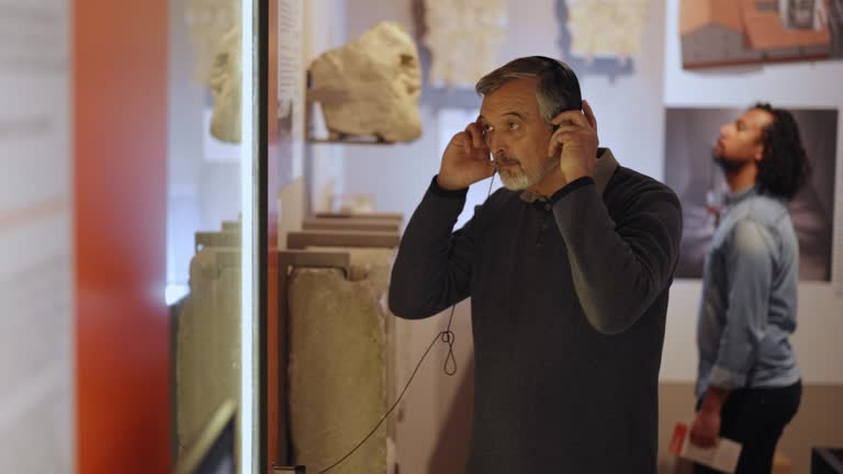 Senior Caucasian Male Putting On Headphones To Listen To The Presentation Of A Displayed Item In A Museum