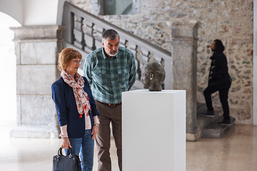 A senior caucasian  woman and a caucasian senior male looking at a bronze bust in a museum. They stopped by the statue and are admiring it. The caucasian senior couple is dressed nicely. The history museum looks old but nead. There is a black man walking up the stairs behind them.