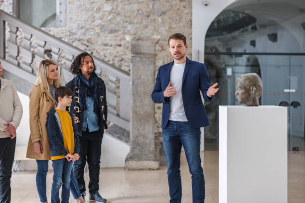 Adult Caucasian Male Museum Tour Guide Giving A Tour Of The Museum To A Diverse Group Of People An adult caucasian male museum tour guide in the middle of a presentation of a bronze statue of a man. He is in the middle of museum tour with a diverse group of museum visitors. He looks confident and professional. He is moving his hands and showing the statue. There is a medieval wall and a staircase behind the people. historical museum stock pictures, royalty-free photos & images