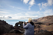 Senior woman toutist taking photos of volcanic rocks on the coast with her smart phone