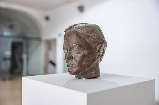 A close up of a bronze head of a man in a museum. The head is put on the white pedestal in the history museum lobby. The lobby is bright and modern.