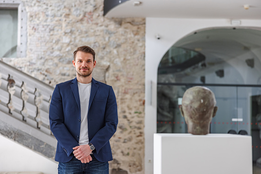 An attractive caucasian male museum employee standing next to a statue of a man's head on a pedestal in a museum. He is looking at the camera. He is dressed professionally. The museum is bright and beautiful with medieval design accents such as a stone wall.