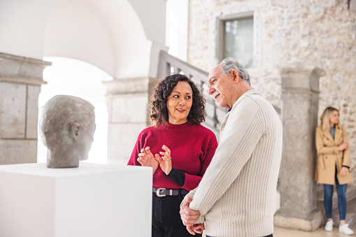 Adult brunette woman having a discussion with a senior caucasian male about the statue in the history museum. They are standing in front of the statue. The male is looking and admiring the statue while the woman is looking at him as she is talking to him. They are standing in a beautiful bright minimalistic history museum with medieval detail such as stone pillars and a staircase.