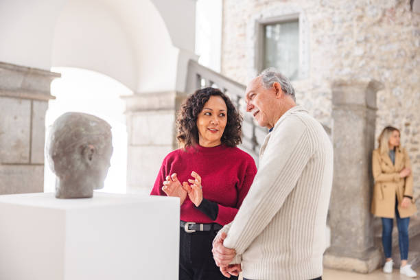 Adult Woman Discussing The Statue With A Senior Caucasian Male In A History Museum Adult brunette woman having a discussion with a senior caucasian male about the statue in the history museum. They are standing in front of the statue. The male is looking and admiring the statue while the woman is looking at him as she is talking to him. They are standing in a beautiful bright minimalistic history museum with medieval detail such as stone pillars and a staircase. historical museum stock pictures, royalty-free photos & images